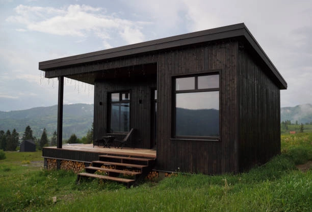 Small black wooden cabin with porch, mountain views, and grassy surroundings. -adu construction