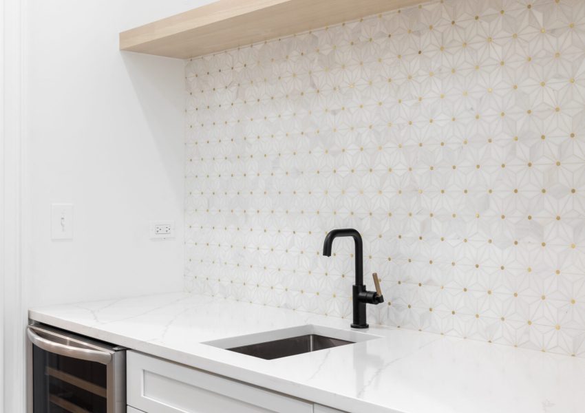 A wet bar with a black faucet, white and gold mosaic tile backsplash, white cabinets, and a stainless steel wine fridge.